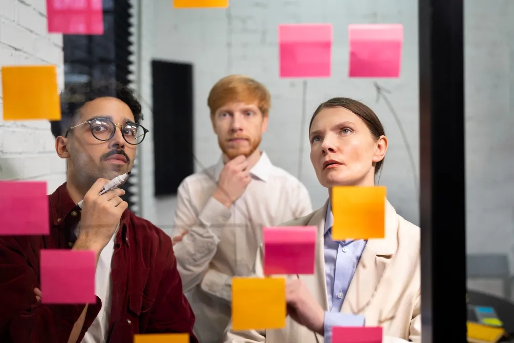 Diverse team analyzing colorful post-it notes on glass wall during meeting.