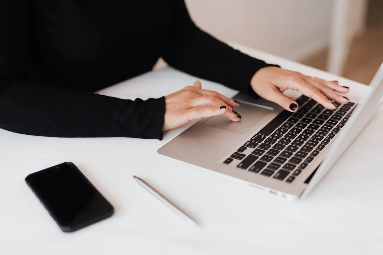 Woman typing on a laptop with a smartphone and pen nearby.